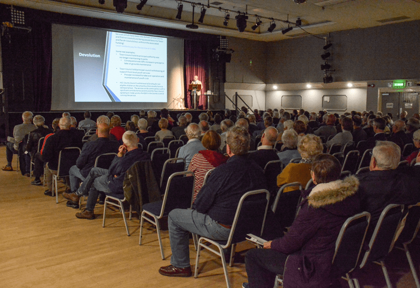 Image of members of the public sat in theatre style seating watching a presentation on a screen on stage